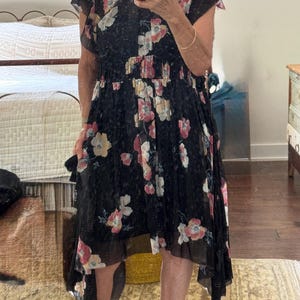 May include: Black floral print dress with a ruffled neckline and asymmetrical hem. The dress features pink and white flowers, a cinched waist, and short sleeves. The fabric appears to be lightweight and flowing.
