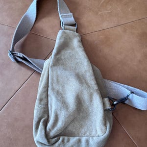 May include: A light tan suede sling bag with an adjustable shoulder strap. The bag has a rounded bottom and a zippered closure. The strap and bag are a matching light tan color. The bag is designed to be worn across the body.