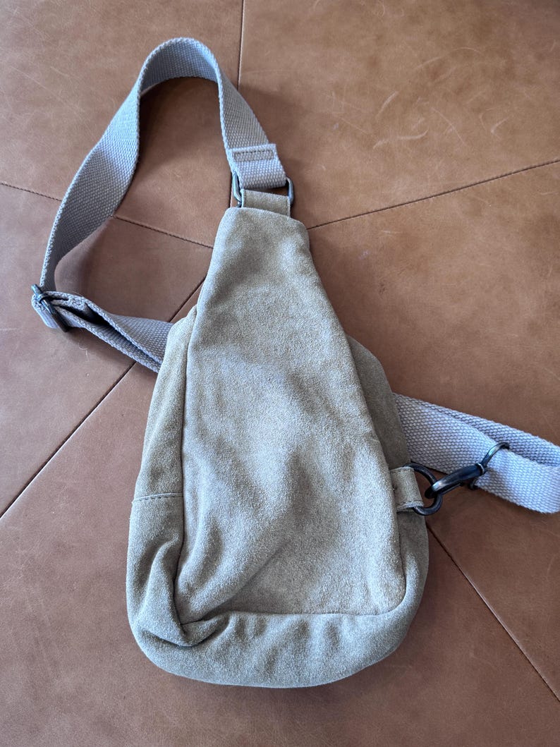 May include: A light tan suede sling bag with an adjustable shoulder strap. The bag has a rounded bottom and a zippered closure. The strap and bag are a matching light tan color. The bag is designed to be worn across the body.