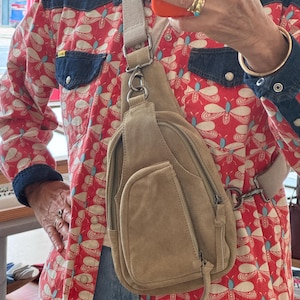 May include: A tan suede sling bag with multiple zippered compartments and a light-colored adjustable strap. The bag is worn across the body. The background includes a red patterned shirt with blue denim accents.