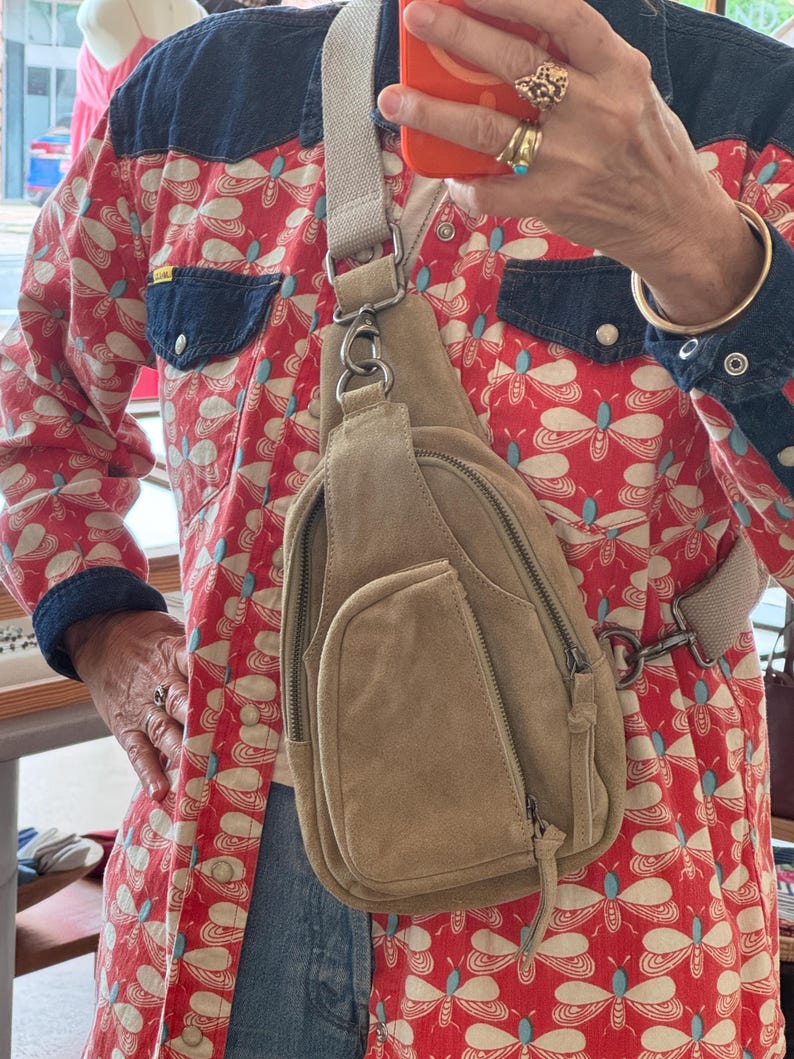 May include: A tan suede sling bag with multiple zippered compartments and a light-colored adjustable strap. The bag is worn across the body. The background includes a red patterned shirt with blue denim accents.