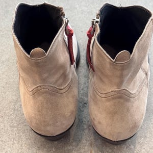 May include: A pair of beige suede ankle boots with zippers and red pull tabs. The boots have a rounded toe and a low heel. The interior is black.