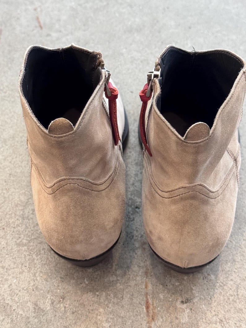 May include: A pair of beige suede ankle boots with zippers and red pull tabs. The boots have a rounded toe and a low heel. The interior is black.