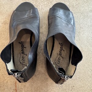 May include: A pair of black leather heeled sandals with a peep toe and zipper closure. The sandals have the text "Free People" written on the inside.