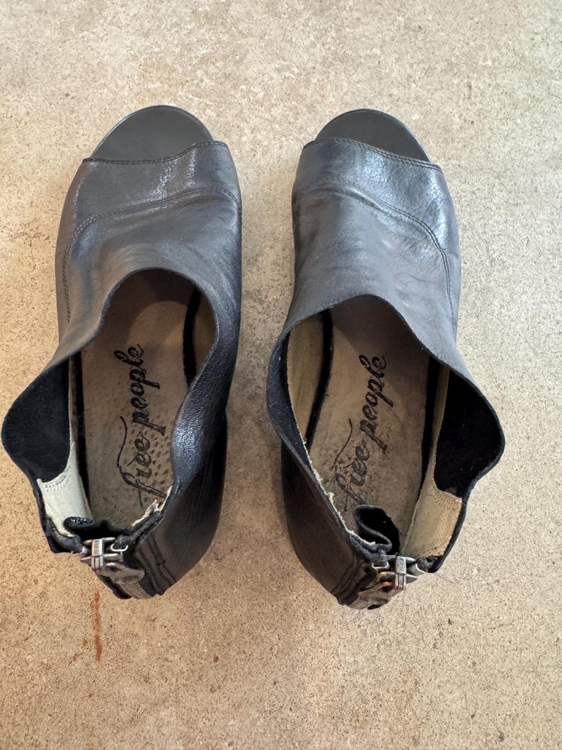 May include: A pair of black leather heeled sandals with a peep toe and zipper closure. The sandals have the text "Free People" written on the inside.