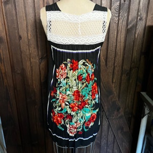 Pu&ograve; includere: Abito nero senza maniche con pannello posteriore in crochet bianco e stampa floreale. L'abito presenta un vivace design floreale in rosso, verde e turchese. Strisce bianche verticali incorniciano il motivo floreale. L'abito &egrave; esposto su un manichino.
