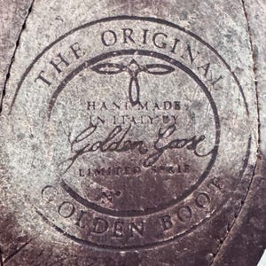 May include: Close-up of a distressed leather surface with a circular emblem. The emblem reads "THE ORIGINAL HAND MADE IN ITALY BY Golden Goose LIMITED EDITION GOLDEN BOOT" in a dark, faded font. The leather has a worn, textured appearance.