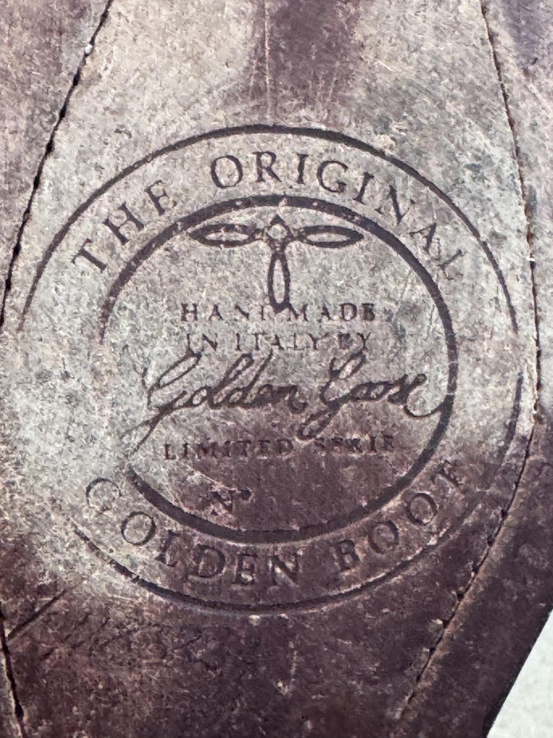 May include: Close-up of a distressed leather surface with a circular emblem. The emblem reads "THE ORIGINAL HAND MADE IN ITALY BY Golden Goose LIMITED EDITION GOLDEN BOOT" in a dark, faded font. The leather has a worn, textured appearance.