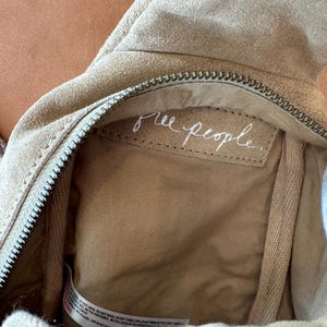 May include: Close-up of a beige suede bag with a zipper closure. The interior features a light brown lining and a tag with the handwritten text "free people". The bag's design suggests a casual, bohemian style.
