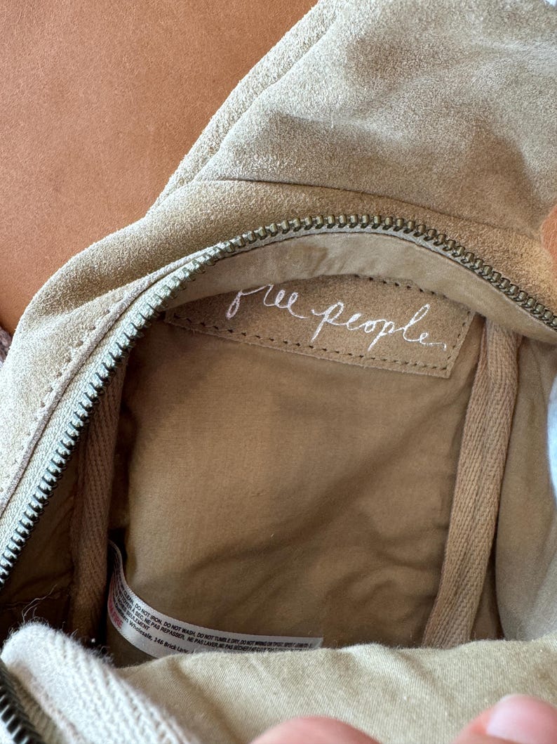 May include: Close-up of a beige suede bag with a zipper closure. The interior features a light brown lining and a tag with the handwritten text "free people". The bag's design suggests a casual, bohemian style.