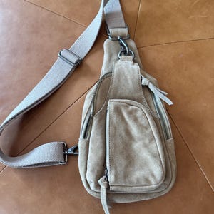 May include: A tan suede sling bag with a long, adjustable canvas strap. The bag has multiple zippered compartments and a small front pocket. The hardware is silver-toned. The bag is designed to be worn across the body.