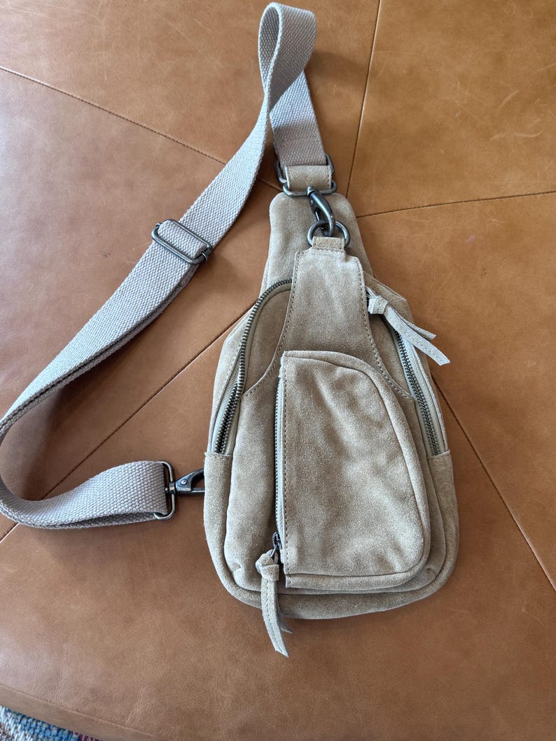 May include: A tan suede sling bag with a long, adjustable canvas strap. The bag has multiple zippered compartments and a small front pocket. The hardware is silver-toned. The bag is designed to be worn across the body.