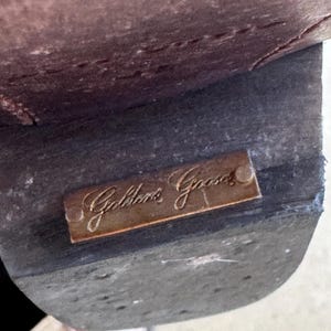 May include: Close-up of a brown leather shoe sole. A small, rectangular, bronze-coloured metal plate is attached to the sole, with the inscription "Golden Goose" in elegant script.