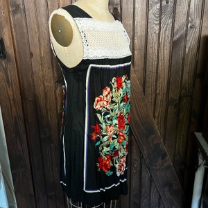 Pu&ograve; includere: Abito nero senza maniche con carr&egrave; in crochet bianco e stampa floreale. L'abito presenta uno scollo quadrato e un colorato motivo floreale con fiori rossi, verdi e bianchi. L'abito &egrave; esposto su un manichino.