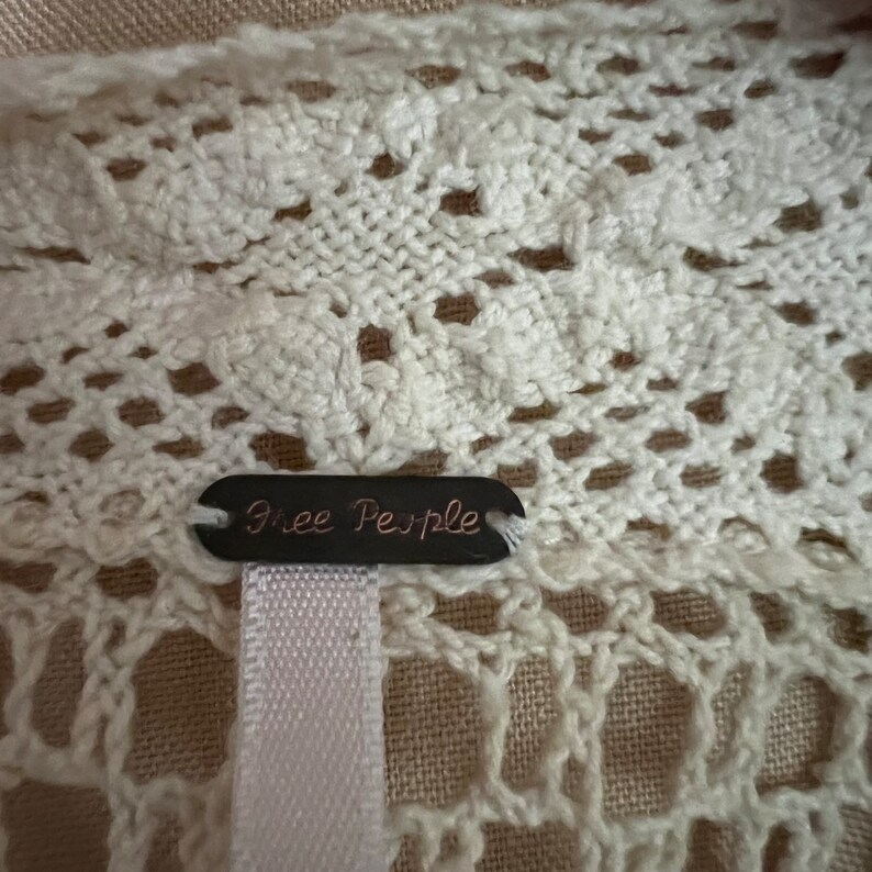 Pu&ograve; includere: Primo piano di un capo in maglia traforata color crema. Il tessuto presenta un delicato motivo a pizzo con piccoli fori e dettagli floreali. Un'etichetta scura con la scritta "Free People" &egrave; attaccata al capo con un nastro bianco.