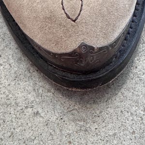 May include: Close-up of a beige suede cowboy boot toe. The toe features a decorative metal cap with an ornate design and a stitched brown pattern. The boot has a black leather sole and a classic Western style.