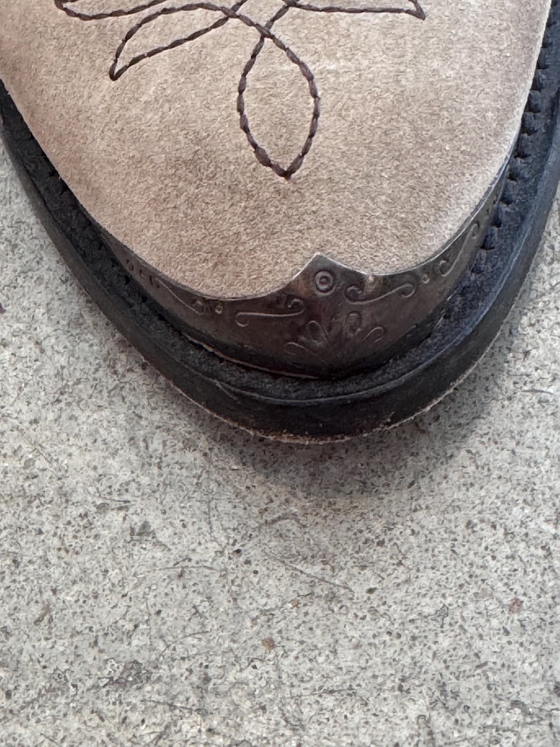 May include: Close-up of a beige suede cowboy boot toe. The toe features a decorative metal cap with an ornate design and a stitched brown pattern. The boot has a black leather sole and a classic Western style.