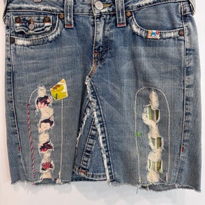 Può includere: Una gonna in denim di lavaggio medio con orlo sfrangiato, sezioni usurate con toppe in tessuto colorato e cuciture bianche visibili. La gonna include una chiusura a bottone, passanti per cintura e cinque tasche con rivetti in metallo.