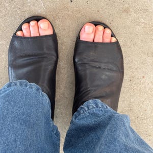 May include: A pair of black leather heeled sandals with open toes. The sandals are worn by a person wearing blue jeans.