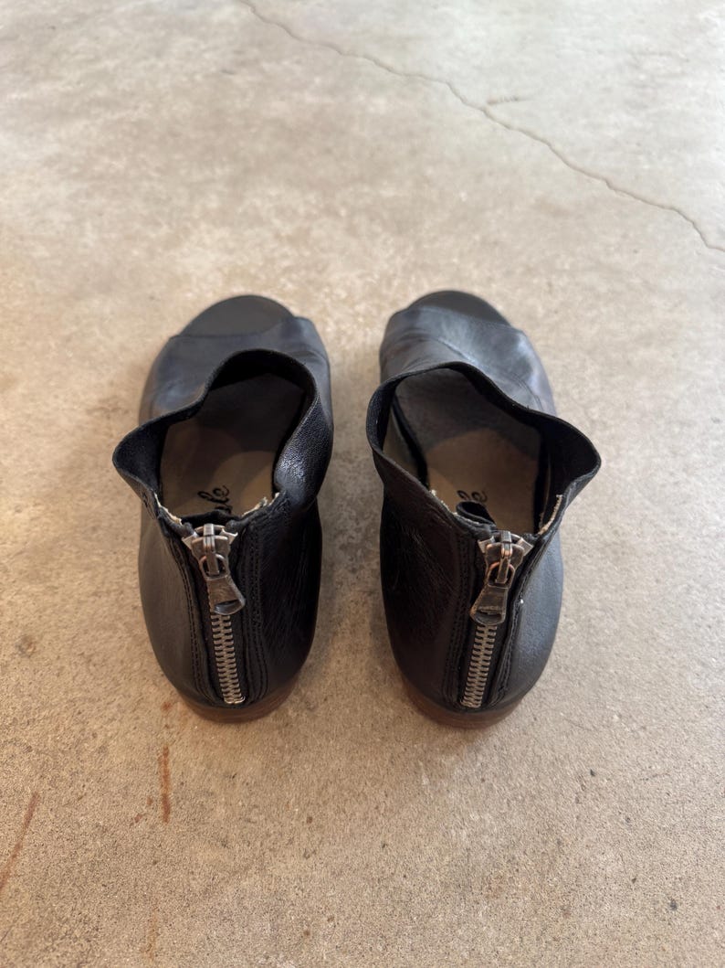May include: A pair of black leather heeled sandals with a zipper detail on the back. The sandals have an open toe and a cut-out design on the side.