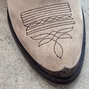 May include: Close-up of a beige suede cowboy boot. The boot features a decorative stitched design in dark brown on the vamp. The toe has a dark brown leather trim and a metal toe cap.