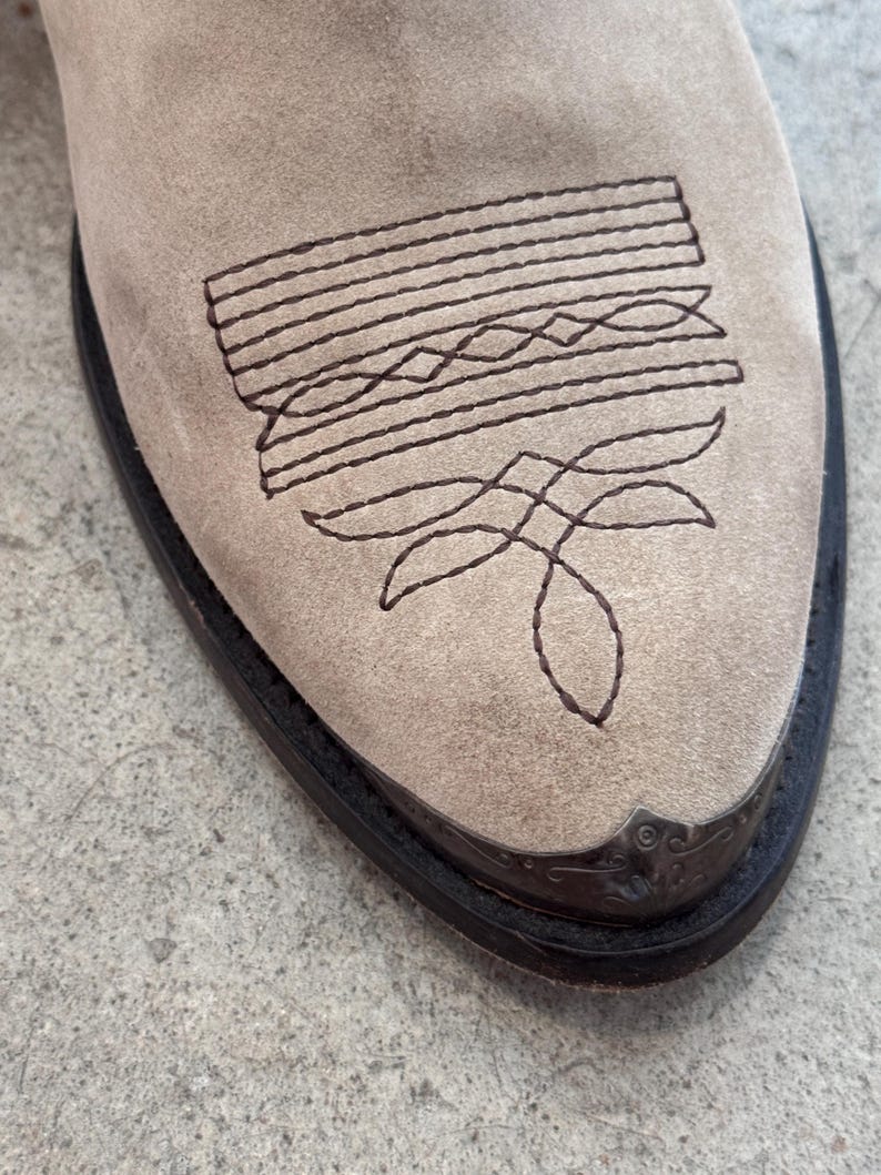 May include: Close-up of a beige suede cowboy boot. The boot features a decorative stitched design in dark brown on the vamp. The toe has a dark brown leather trim and a metal toe cap.