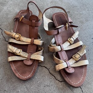 May include: A pair of flat sandals featuring brown leather footbeds and straps, accented with metallic gold leather straps across the front. Each sandal includes multiple adjustable gold-tone buckles and a heel strap with a buckle closure.