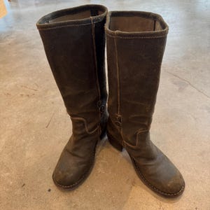May include: A pair of dark brown suede knee-high boots. The boots have a worn, distressed appearance, with visible seams and a zipper detail on the side. The soles are a darker shade of brown.