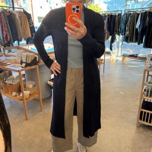 May include: A long, black cardigan with a draped front. The cardigan is worn over a light blue top and tan pants. The image is taken in a clothing store, with racks of clothing visible in the background.