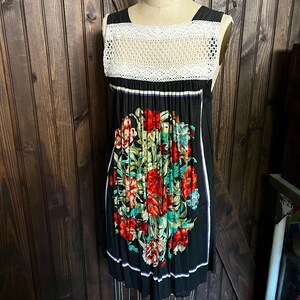 Pu&ograve; includere: Abito nero senza maniche con corpetto in crochet bianco e stampa floreale. L'abito presenta uno scollo quadrato e una gonna a pieghe. La stampa floreale comprende fiori rossi, verdi e blu. L'abito &egrave; esposto su un manichino.