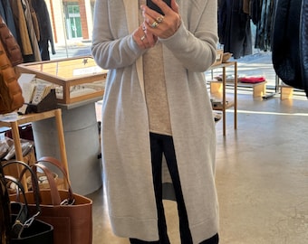 May include: A light gray long cardigan with a hood and a contrasting beige trim. The cardigan is open in the front and worn over a beige sweater and dark pants. The image is taken in a store setting with other clothing racks visible.