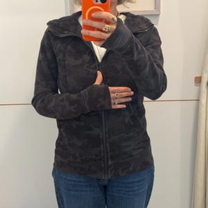 May include: A black and gray camouflage print zip-up hoodie with a high collar and thumbholes.