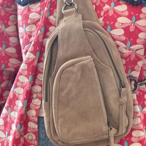 May include: A tan suede sling bag with multiple zippered compartments and a small front pocket. The bag features a silver-tone metal chain and hardware. The background includes a red patterned shirt with blue denim accents.