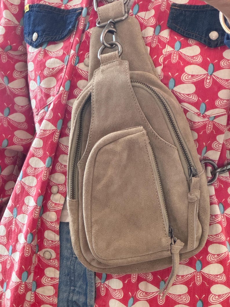 May include: A tan suede sling bag with multiple zippered compartments and a small front pocket. The bag features a silver-tone metal chain and hardware. The background includes a red patterned shirt with blue denim accents.