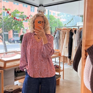 May include: A woman wearing a pink and white patterned blouse with a v-neck and long sleeves. She is also wearing dark blue pants.