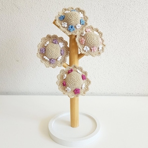 May include: Four crocheted hats with floral accents displayed on a wooden stand. The hats are in shades of beige, pink, blue, and purple.