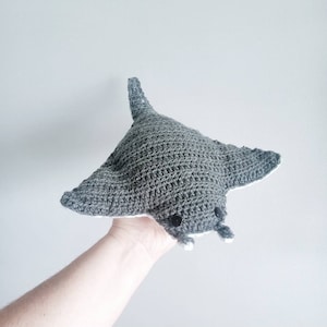 Puede incluir: Un animal de peluche de manta raya de crochet gris con detalles blancos alrededor de los bordes y ojos negros.