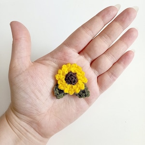 May include: A small crocheted sunflower with yellow petals and a brown center, with two green leaves attached.