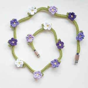 Puede incluir: Un cable de carga de teléfono de crochet verde con flores moradas y blancas adjuntas.