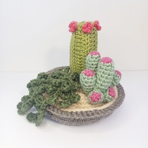 May include: A crocheted cactus plant with pink flowers in a gray pot. The cactus is green and has a light green base. The pot is filled with a light brown material.