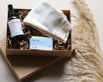 May include: A brown gift box filled with brown paper shreds, containing a white baby onesie, a bottle of stretch mark oil, and a bar of baby soap. The box is labeled "Arlio".