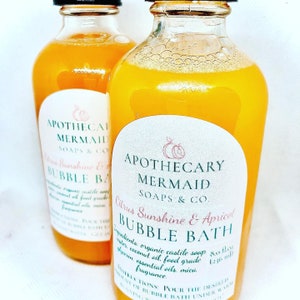 May include: Two glass bottles of citrus sunshine and apricot bubble bath from Apothecary Mermaid Soaps & Co. The bottles are filled with a yellow liquid and have white labels with black text. The label includes the ingredients: organic castile soap, water, coconut oil, food grade glycerin, essential oils, mica, and fragrance. The label also includes instructions for use: Pour the desired amount of bubble bath under warm running water.