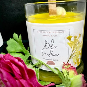 May include: A yellow candle in a glass jar with a wooden wick. The label reads "Apothecary Mermaid Soaps & Co. Boho Sunshine Hand Poured Candle 13 Ounces".