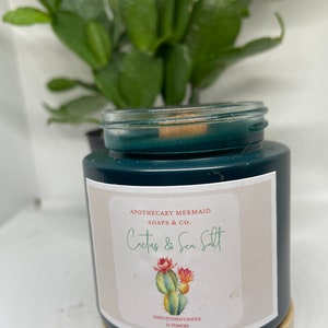 May include: A dark green glass candle jar with a white label that reads "APOTHECARY MERMAID SOAPS & CO. Cactus & Sea Salt" and a hand-drawn illustration of a cactus with pink flowers. The label also states "HAND POURED CANDLE 13 OUNCES".