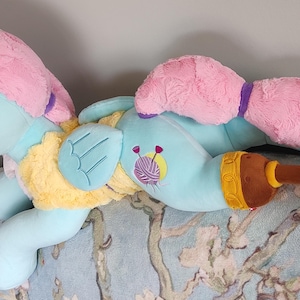 May include: A plush pony toy in a lying pose, with a light blue body, pink mane and tail, and yellow and brown details. The toy has a cartoon design with embroidered details, including a yarn ball. The pony is resting on a floral patterned surface.
