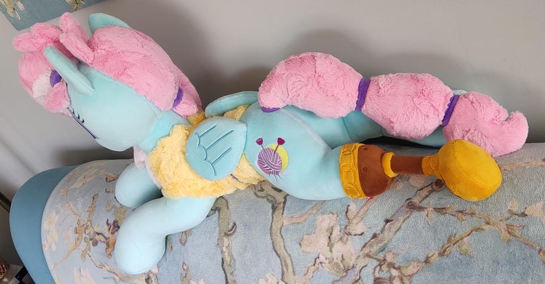 May include: A plush pony toy in a lying pose, with a light blue body, pink mane and tail, and yellow and brown details. The toy has a cartoon design with embroidered details, including a yarn ball. The pony is resting on a floral patterned surface.