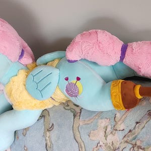 May include: A plush toy pony in a lying position, featuring a light blue body, pink mane and tail, and yellow accents. The toy has a brown and yellow accessory. The pony has a yarn and needle design on its body.
