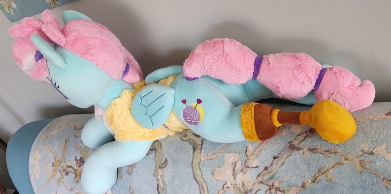 May include: A plush toy pony in a lying position, featuring a light blue body, pink mane and tail, and yellow accents. The toy has a brown and yellow accessory. The pony has a yarn and needle design on its body.