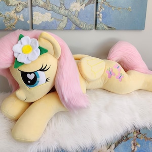 Puede incluir: Un juguete de peluche de un pony rosa y amarillo con una flor blanca en la cabeza. El pony est&aacute; acostado sobre una superficie blanca y esponjosa.