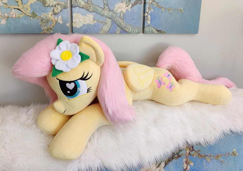Puede incluir: Un juguete de peluche de un pony rosa y amarillo con una flor blanca en la cabeza. El pony est&aacute; acostado sobre una superficie blanca y esponjosa.